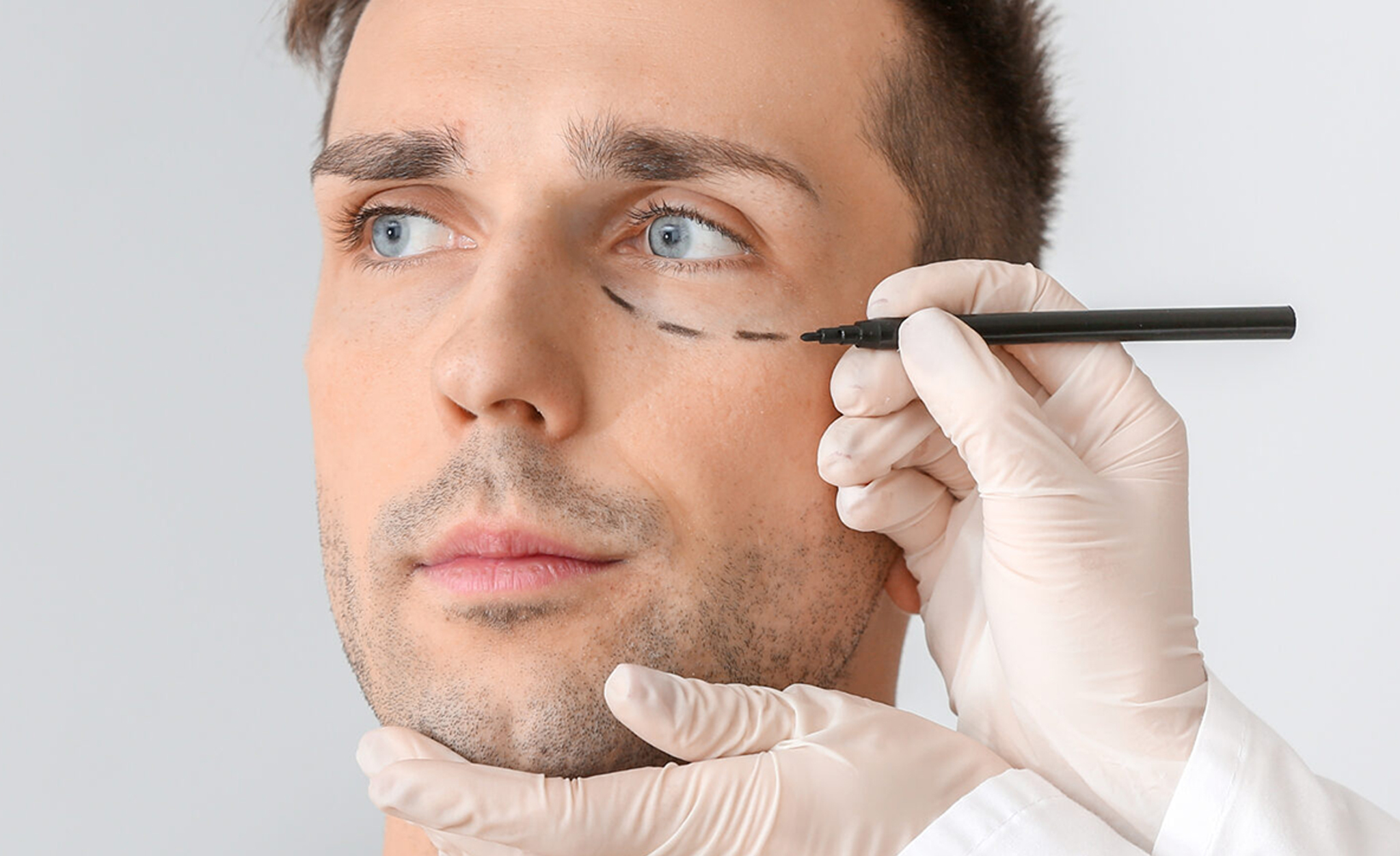 Plastic surgeon applying marking on male face against light background